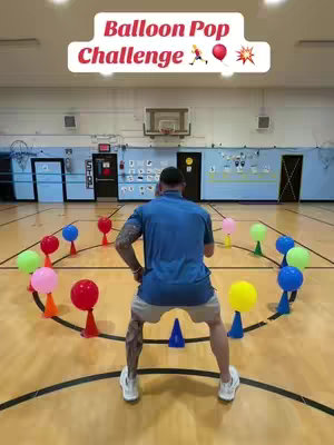 A man in a blue shirt and khaki shorts stands on a basketball court, facing away from the camera. He appears to be interacting with a circle of colorful balloons set on cones. Above him is a text overlay that says 'Balloon Pop Challenge' in a bubble letter style.