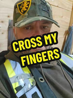 A selfie of a man wearing a camouflage baseball cap, a work vest, and safety glasses, with the text overlay 'CROSS MY FINGERS' in yellow. The background is a wooden surface.
