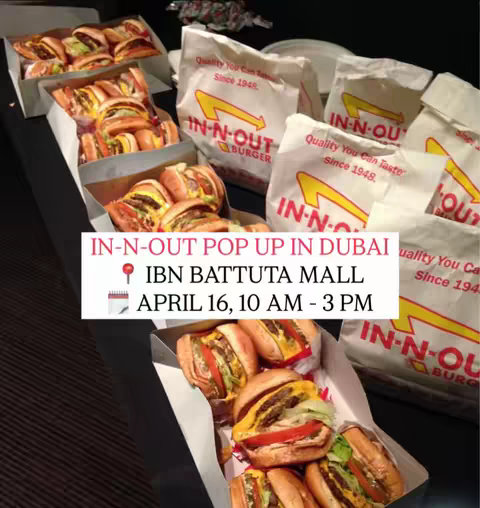 The image shows several boxes and bags of In-N-Out burgers and packaging on a dark surface. Text overlays provide details about an upcoming pop-up event in Dubai, including the location and date/time details.
