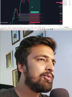 A man with a beard and dark hair is looking to the right. Behind him is a monitor with a trading-related chart displayed. Charts show green and red candle sticks. The background is a room with square-framed imagery on the wall. There is a microphone on the table in front of him.