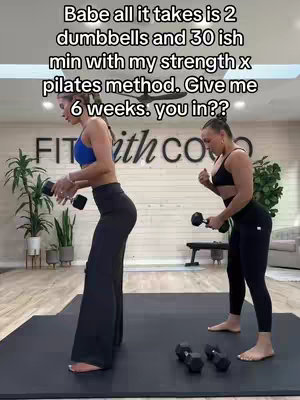 Two women are in a gym setting, engaged in what appears to be a strength and pilates workout. One woman, on the left, is wearing black pants and a blue top, holding a dumbbell in each hand, and in a forward-leaning position. The second woman, to the right, is wearing a black sports bra and leggings, working out in a similar pose with dumbbells. The background includes exercise equipment, a white brick wall, and potted plants, indicating they are indoors at a gym.