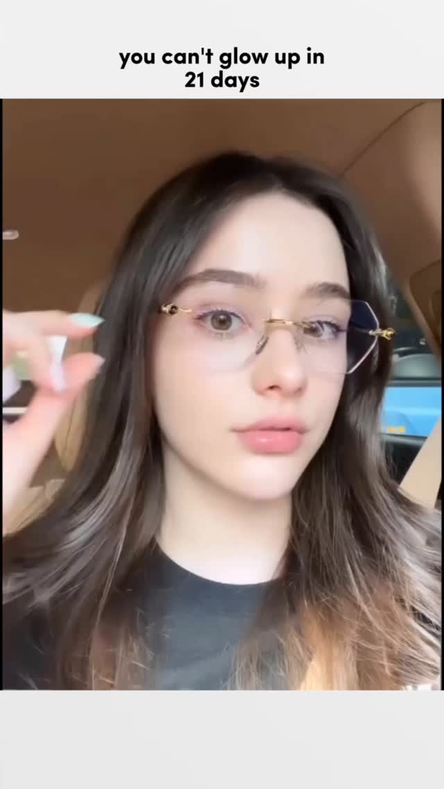 A young woman with long brown hair wearing glasses sits in a car. The image is a close-up of her face, and she is holding up her finger to adjust her glasses. The background is blurred, suggesting a car interior. The overall composition is a selfie style.