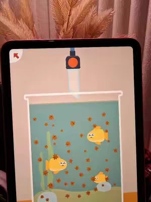 The image shows a tablet displaying a mobile game. The game features a fish tank with yellow cartoon fish, brown leaves, and green plants inside. A cleaning tool is positioned above the tank. The background is a soft gradient of pink and brown, with some out-of-focus leafy elements.