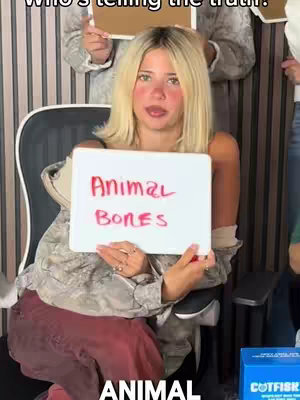 A woman with long blonde hair sits in an office chair holding a white board reading "Animal Bones" in red marker. Other individuals are partially visible in the background, each holding up a similar white board. The overall setting appears to be a game or guessing game, with a blue box labeled "CATFISH" partially visible in the bottom right.