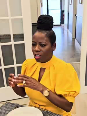 A woman with dark hair pulled into a bun is talking in a well-lit room. She wears a yellow dress and a watch. A table setting is visible in the foreground, and a white door and hallway are in the background.