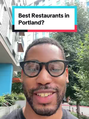 A medium shot of a Black man with short black hair and black glasses. He is smiling and looking at the camera. Behind him, there's a building and some green trees.