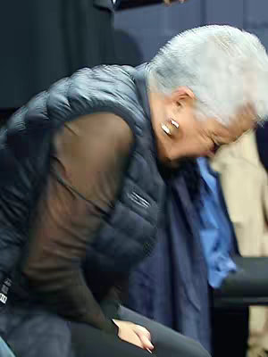 A woman with short grey hair is bent over, likely in a seated position, with a partially visible black vest and mesh top. The image is blurry, giving a sense of movement or intimacy. Darker backgrounds indicate a possible indoor setting.