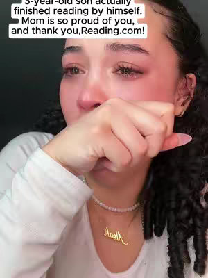 A close-up shot of a person, presumably a mother, crying. She is holding her hand up to her face, partially obscuring it, which is a gesture of distress and sadness. Her face is the primary focus, with tears visibly streaming down her face. The background is blurred and out of focus, but it seems to be indoors, possibly a room with soft lighting. The person is wearing a white top, and a gold necklace with a name pendant hangs around her neck. The overall composition is intimate and emotional, emphasizing the woman’s vulnerability and her display of grief.