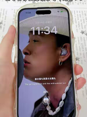 The image features a close-up shot of a smartphone displaying a lock screen with a picture of a man singing into a microphone. The man is wearing a hat and an earring. The lock screen displays the time and battery percentage, along with a Japanese caption. The phone is held by a hand with a clear phone case.