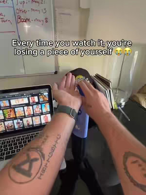 The image shows a first-person perspective with hands, with the focus being on a laptop screen displaying images of adult content. A hand is holding an object, possibly a blue cloth, in front of the screen. The background includes a whiteboard with writing, a window, and a desk with items on it. The image is framed to convey the creator's body at work while drawing your attention to the content on the laptop screen.