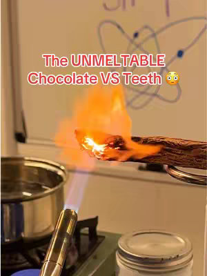 The image shows a piece of chocolate being set on fire by a butane torch. The chocolate is held with tongs. In the background, there is a whiteboard with scientific diagrams and writing. In the foreground, a stainless steel pot and a jar are visible.