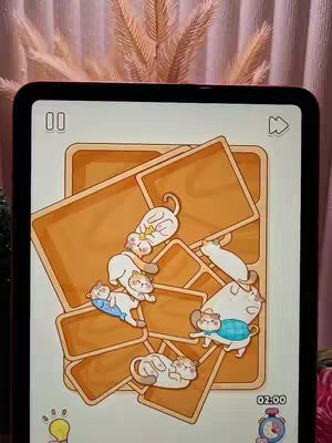 An iPad screen is displaying a puzzle game with various orange-colored rectangular shapes and playful cartoon cat illustrations. The cats are depicted in varied poses and are wearing clothes like a blue shirt and an orange collar. The layout suggests the game interface, focusing on problem-solving and visual appeal.
