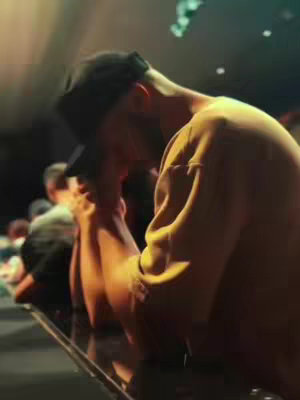 The cover image shows a man wearing a black cap, with his head bowed and his hands clasped in what seems to be prayer or contemplation. He is wearing a yellow sweatshirt. The background is blurred and appears to be a stage or performance setting, with some people visible in the background.