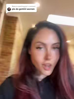 A selfie-style video showing a woman with long, dark hair with red highlights looking directly at the camera and speaking. The background is slightly blurred but appears to be indoors with warm-toned walls.