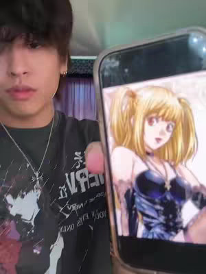 A young man is holding up his phone, which displays an anime character on the screen. The man is centered in the frame, looking towards the viewer with a slight frown. The background is blurred, while the man's upper body and the phone are in sharp focus. The phone is also in sharp focus, with the anime character Misa Amane from Death Note. The man is wearing a black t-shirt with white graphics, and he has a silver necklace.