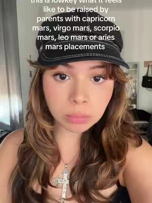 A young woman with long brown hair and a black cap is the focus. She is wearing a necklace with a cross pendant. The background includes parts of a room, with a mirror, a black handbag and other items. The text overlays on top of the image.