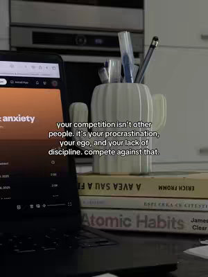 A laptop displaying a music streaming app is open atop a desk, with a stack of books, a cactus-shaped pen holder filled with pens, and other stationary. The background is blurred, suggesting a workspace.