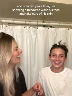 A candid shot shows a mother with long blonde hair and her son smiling in a bathroom. The son has a white substance on his face, looking toward the camera. The background features a white shower curtain.