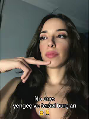 A woman with long, wavy brown hair is centered in the frame, looking at the camera. She has a slight smile and is touching her chin with a finger, and there is white text on the bottom of the image. The background has dark tones.