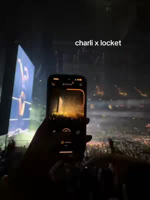 A person is holding a phone up in front of them, showing a concert stage and crowd through the phone's screen. The phone screen displays a concert scene bathed in warm lighting. The image conveys a sense of being at a Charli XCX concert. The camera is angled from a slightly low perspective, with the hand and phone taking up the majority of the frame's foreground.