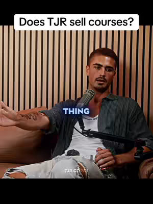 The image shows a man wearing a dark blue shirt sitting on a sofa with vertical wooden paneling in the background. The man has a tattoo is visible on his left arm. A microphone is positioned in front of him. White text at the top says "Does TJR sell courses?" and blue text at the center says "THING".