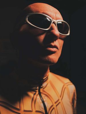 A close-up shot of a person wearing high-quality, futuristic white framed sunglasses against a dark background. The subject has smooth skin and a bald head. They are wearing an intricate, geometric pattern top, which is lit with a dramatic lighting scheme to create depth and visual interest.