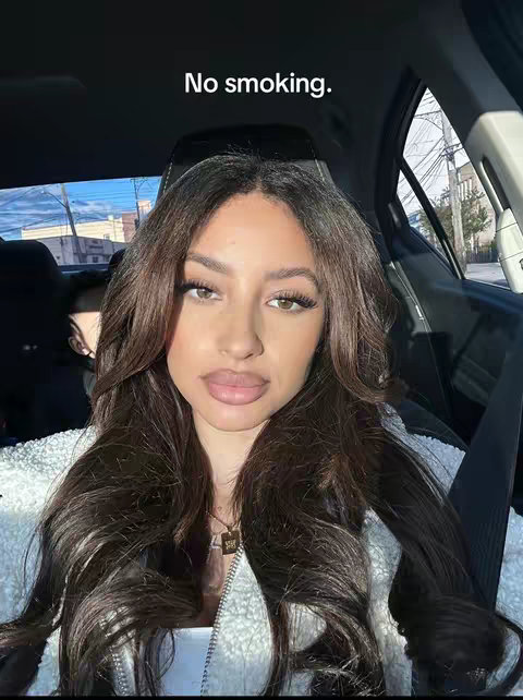 A selfie showing a woman sitting in a car, with her face taking up most of the frame. She has long, dark brown hair and is wearing a white fluffy jacket. The text 'No smoking' is overlaid on the top. The background shows a street scene through the car window.