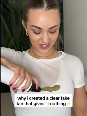 A young woman with blonde hair is shown from the chest up, angled slightly downward. She wears a white t-shirt and a gold necklace. Her expression is pleasant, and she appears to be smiling. She is holding a white bottle and squeezing out a dark brown substance, perhaps self-tanner, which is pooling on her shirt. Text at the bottom of the image claims the product is "a clear fake tan". The background is neutral with a suggestion of indoor greenery.