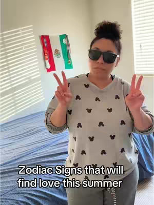 A woman in a gray Mickey Mouse print sweatshirt and sunglasses makes a peace sign with both hands. Behind her is a bedroom with a Mexican flag hanging on the wall and a bed with blue sheets.