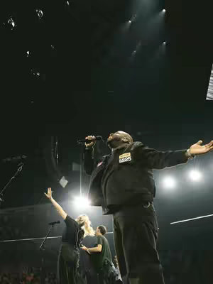 A live music stage with multiple performers. The main subject, a man in a black jacket and pants, is singing into a microphone with arms outstretched. Another two performers are standing stage left, one raising their arms and the other appears to be singing.