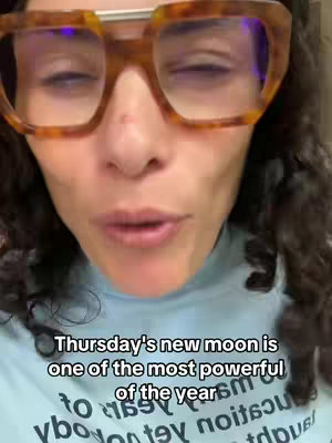 A close-up shot featuring the upper body of a woman with curly hair and large, patterned glasses. Her eyes are closed, and she is speaking. Text in a stylized font is overlaid near the bottom of the frame, describing a powerful new moon.