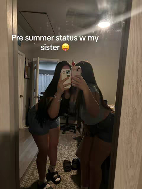 Two young women are reflected in a mirror, taking a selfie. The mirror is large and framed, showing a room with neutral walls and a textured carpet. Both women are holding phones and taking the photo, with their faces partially obscured by the phone screens. The lighting appears to be natural or soft, with the overall color scheme being muted. Text is overlaid near the top.