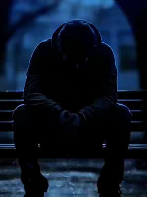 A silhouette of a person sitting on a bench, facing downward. The person is wearing a hooded jacket, obscuring the face. The scene is bathed in a cool, blue light, suggesting a night or outdoor setting. The composition emphasizes the figure's isolation and introspection.