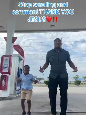 A medium shot at a gas station featuring two people. The foreground shows a young person and an adult. The young person, wearing a light blue shirt and khaki shorts, stands on the left side of the frame, while the adult, dressed in a dark green shirt and black pants, stands on the right. Both have their arms spread apart as if dancing. The background includes gas station pumps and a cloudy sky.