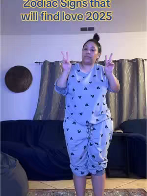 A woman stands indoors, wearing pajamas with heart-shaped patterns. She holds up two fingers on each hand in a peace sign. The background includes a dark blue couch, curtains, and a round decorative object on the wall.