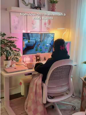 Scene shows a person sitting at a desk, viewed from behind, with a computer displaying a video game. The desk has a clean, organized setup. A plant and decorative items are on the desk, and a blanket is draped over the chair. The color palette is warm and inviting, with soft lighting.