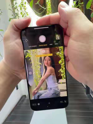 The cover image shows a person holding an iPhone in front of them, capturing a photo of a young woman. The woman is sitting in a well-lit, mostly white space. The phone screen displays the camera interface, with the subject centered in the frame. The hands holding the phone are visible, and the composition suggests a photo is being taken. There is greenery in the background and on the phone frame.