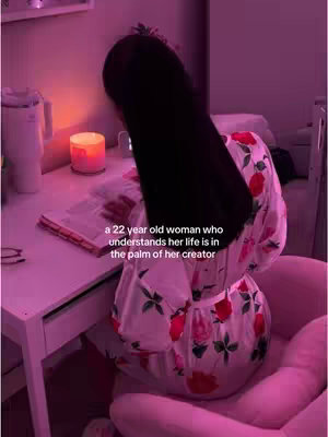 The image captures a person from behind, seated at a desk with a soft pink glow from a nearby candle. The person is wearing a flowery robe and has long, dark hair that drapes over their back and shoulders obscuring the face. On the desk is a book and various items contributing to a setting that conveys a sense of intimacy and introspection.