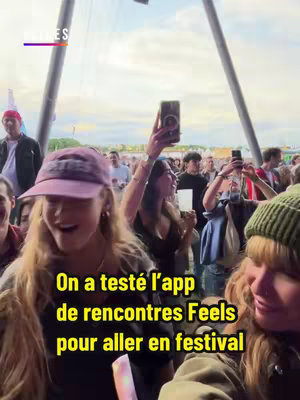A selfie-style shot taken at a festival. In the immediate foreground, two young women are visible; one is wearing a hat and the other is holding a phone up in the air. Many people are in the background.
