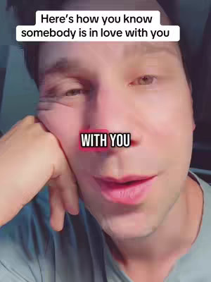 A close-up shot of a fair-skinned man with brown hair. He is looking towards the upper right of the frame, and his left arm is resting on a surface, with his hand supporting his head. The background is blurred, suggesting the focus is on him. There are two text overlays. The top one reads in a white bold sans-serif font: 'Here's how you know somebody is in love with you'. Beneath this, in a pink and black outlined text box (with a black shadow) is the text: 'WITH YOU'