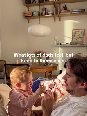 A warm, bright interior shot featuring a father and his baby. The father, with dark hair and a beard, is seated with the baby on his lap. The baby is reaching for a pair of sunglasses held by the father, and both are smiling. The background includes soft, neutral tones from the sofa, walls, and floating shelves, creating a cozy atmosphere. A large spherical light fixture hangs from the ceiling, complemented by a vase of flowers to the left. The text overlay is centered at the top, against the backdrop.