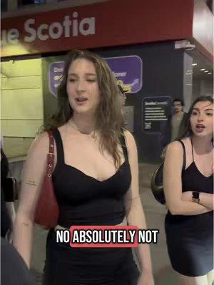 A woman in a black top and skirt is being interviewed on a street at night. Behind her is a wall with a business sign on it.