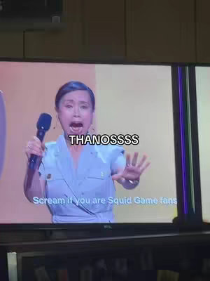 A woman on a large screen is holding a microphone and yelling with an open mouth. Text overlay says "THANOSSSSS" on top, with "Scream if you are Squid Game fans" below.