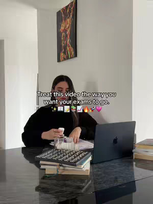 A woman sits at a dark countertop with a laptop, books, and papers. The background includes a framed abstract artwork and white walls. The video overlay shows the message "Treat this video the way you want your exams to go." with emojis.