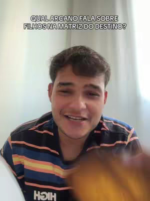 A young man with short brown hair and a small ear piercing is positioned in front of a light-colored wall. He's wearing a black, blue, orange, and white striped shirt while smiling at the camera. Above him, the text reads, "QUAL ARCANO FALA SOBRE FILHOS NA MATRIZ DO DESTINO?"