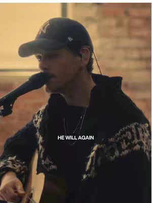 A man wearing a backwards baseball cap and a black sweater plays a guitar and sings into a microphone. The background is blurred.