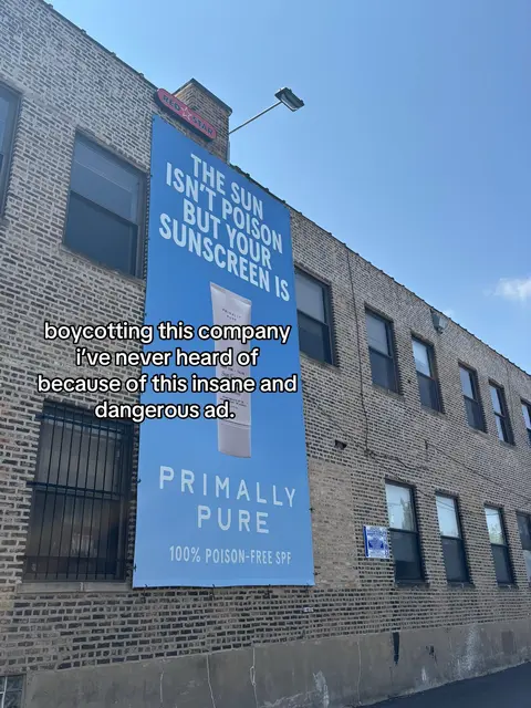 A daytime shot of a brick building with a large blue banner hanging on the side. The banner has white text that reads: “THE SUN ISN'T POISON BUT YOUR SUNSCREEN IS”. Below, in smaller text, it promotes “PRIMALLY PURE 100% POISON-FREE SPF”. Below the banner is a line of handwritten black text on a white background. The sky above is clear and blue.