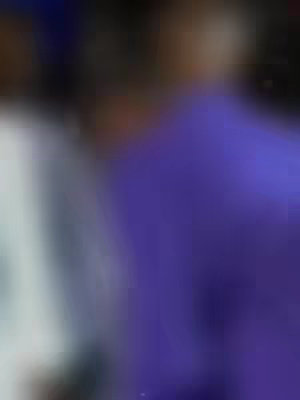 The image is heavily blurred, with a blurry figure wearing a purple garment on the right, partially obscured by another figure in white or light-colored clothing on the left. The background is dark and indistinct.