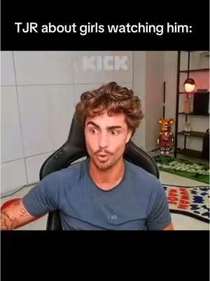 A man with curly brown hair and a mustache is seated in a gaming chair, looking at the camera with a surprised expression. The background includes a KICK logo, a shelf with items like toy figures and a lamp. The man is wearing a blue t-shirt.