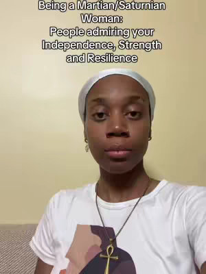 A medium shot features a dark-skinned woman with a white head covering and a white t-shirt. Text overlays at the top state, 'Being a Martian/Saturnian Woman: People admiring your Independence, Strength and Resilience.' The background is a plain, light beige wall. She looks directly at the camera with a neutral expression. A golden necklace with an Ankh pendant is visible.